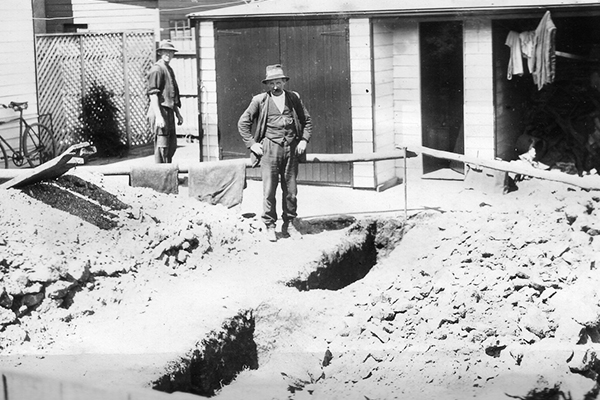 Photograph of early residential sewer connection, worker admiring the trench.