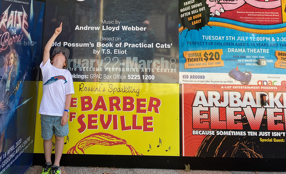 Image of boy pointing upwards to a scavenger hunt clue. He is pictured in front of theatre posters at Geelong Arts Centre.  
